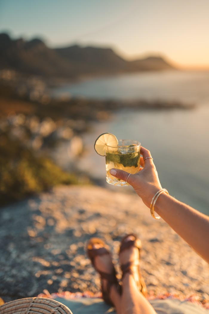 Home A hand holding a mojito cocktail against a scenic ocean view at sunset in Cape Town, South Africa.